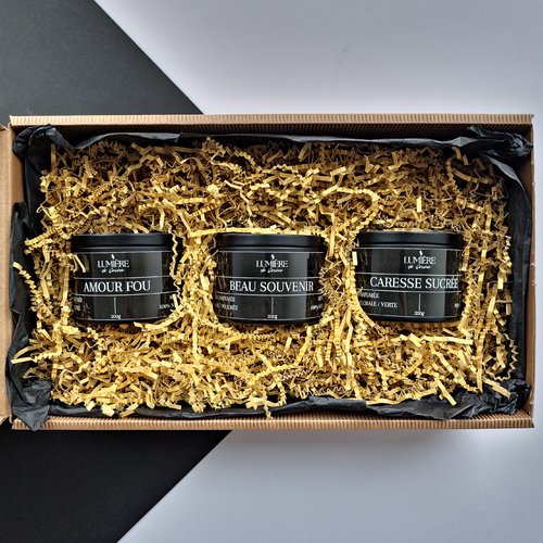 L'image montre une boîte cadeau contenant trois pots noirs avec du texte blanc, chacun étiqueté avec des noms différents. Les pots sont placés sur un lit de papier doré déchiqueté. Les étiquettes sur les pots indiquent :
"AMOUR FOU" - 200g
"BEAU SOUVENIR" - 200g
"CARESSE SUCRÉE" - 200g