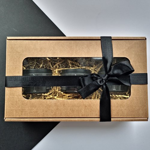 L'image montre une boîte cadeau en carton ondulé brun avec une fenêtre transparente sur le dessus. À l'intérieur de la boîte, il y a trois pots noirs partiellement visibles à travers la fenêtre. La boîte est remplie de papier déchiqueté pour amortir les chocs. Elle est également nouée avec un ruban noir attaché en boucle sur le côté droit. Le contraste entre le carton brun, le ruban noir et les pots noirs à l'intérieur rend la présentation visuellement attrayante.
