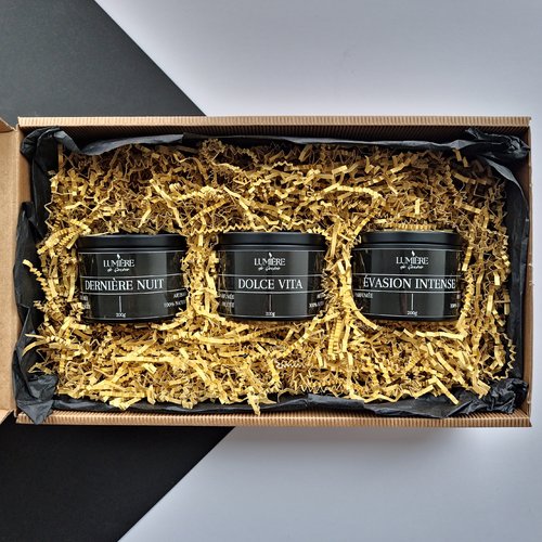 L'image montre une boîte cadeau contenant trois bougies noires nichées dans du papier déchiqueté jaune. Les bougies sont étiquetées "DERNIÈRE NUIT," "DOLCE VITA," et "ÉVASION INTENSE." Chaque bougie porte la marque "LUMIÈRE de Genève" et pèse 200g. L'emballage est soigné et esthétiquement agréable, ce qui en fait une option cadeau attrayante.