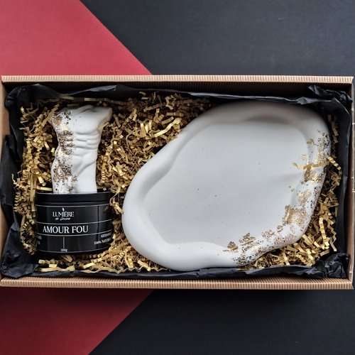 L'image montre une boîte cadeau contenant une bougie et un objet décoratif. La bougie, étiquetée "LUMIÈRE de Genève" et AMOUR FOU", est placée à gauche et ornée d'un design sculpté en forme de torse. À droite, se trouve un objet décoratif blanc à la forme irrégulière avec des accents dorés. Les articles sont nichés dans du papier déchiqueté pour la protection et la présentation. La boîte est en carton avec un fond noir et rouge, suggérant un ensemble cadeau luxueux et artistique.