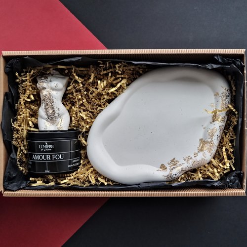 L'image montre une boîte cadeau contenant trois éléments : une sculpture blanche représentant un torse, un contenant cylindrique noir étiqueté "LUMIÈRE de Genève" et "AMOUR FOU", et un grand objet blanc de forme irrégulière avec des accents dorés. Les objets sont nichés dans du papier d'emballage déchiqueté. La sculpture et le contenant sont positionnés sur le côté gauche, tandis que le grand objet blanc est à droite. L'arrière-plan de la boîte est noir et la boîte elle-même est en carton.