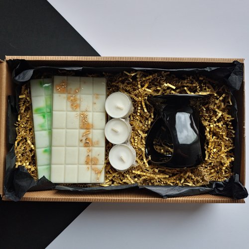 L'image montre une boîte cadeau contenant plusieurs articles disposés sur un lit de papier doré déchiqueté. Les articles comprennent :
Un brûleur d'huiles en céramique noire.
Trois bougies chauffe-plat blanches.
Deux barres de fondants de cire, l'une avec des accents verts et l'autre avec des flocons dorés.
Cette composition présente un coffret cadeau soigneusement conçu, probablement destiné à l'aromathérapie ou aux parfums d'intérieur.