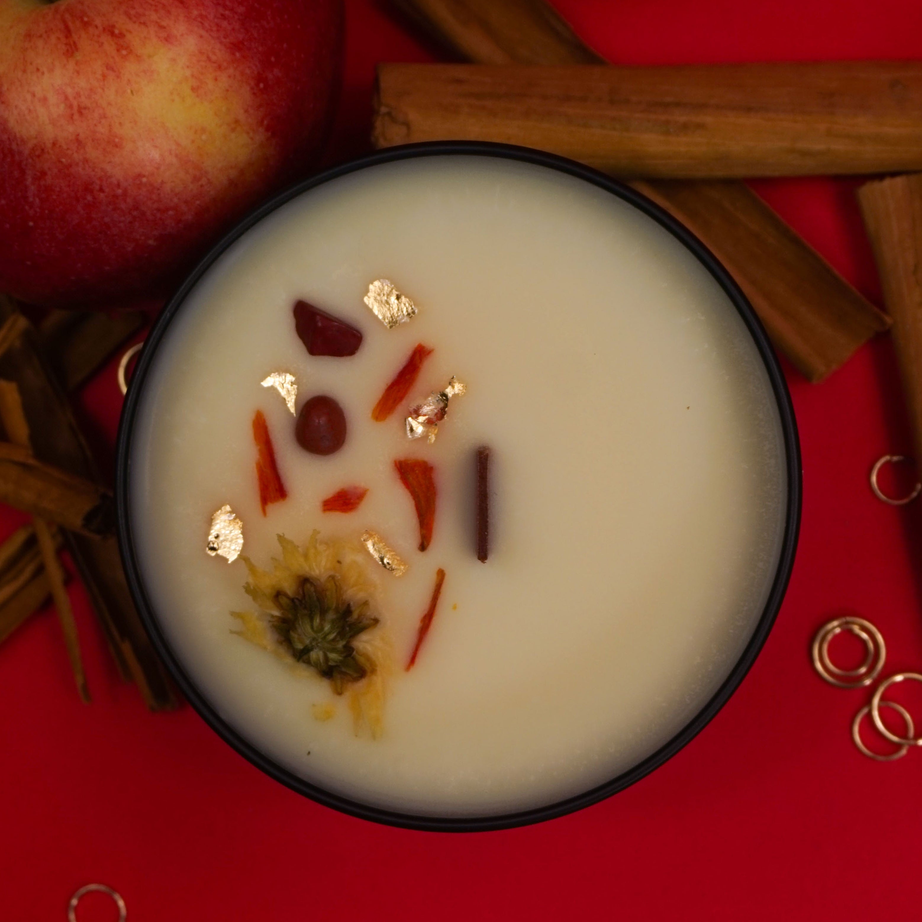Photographie d'une bougie parfumée dans un contenant noir.
La surface de la bougie est décorée de morceaux séchés de pomme et de cannelle. Autour de la bougie, des pommes rouges et des bâtons de cannelle sont posés sur un fond rouge vif, créant une ambiance chaleureuse et épicée."