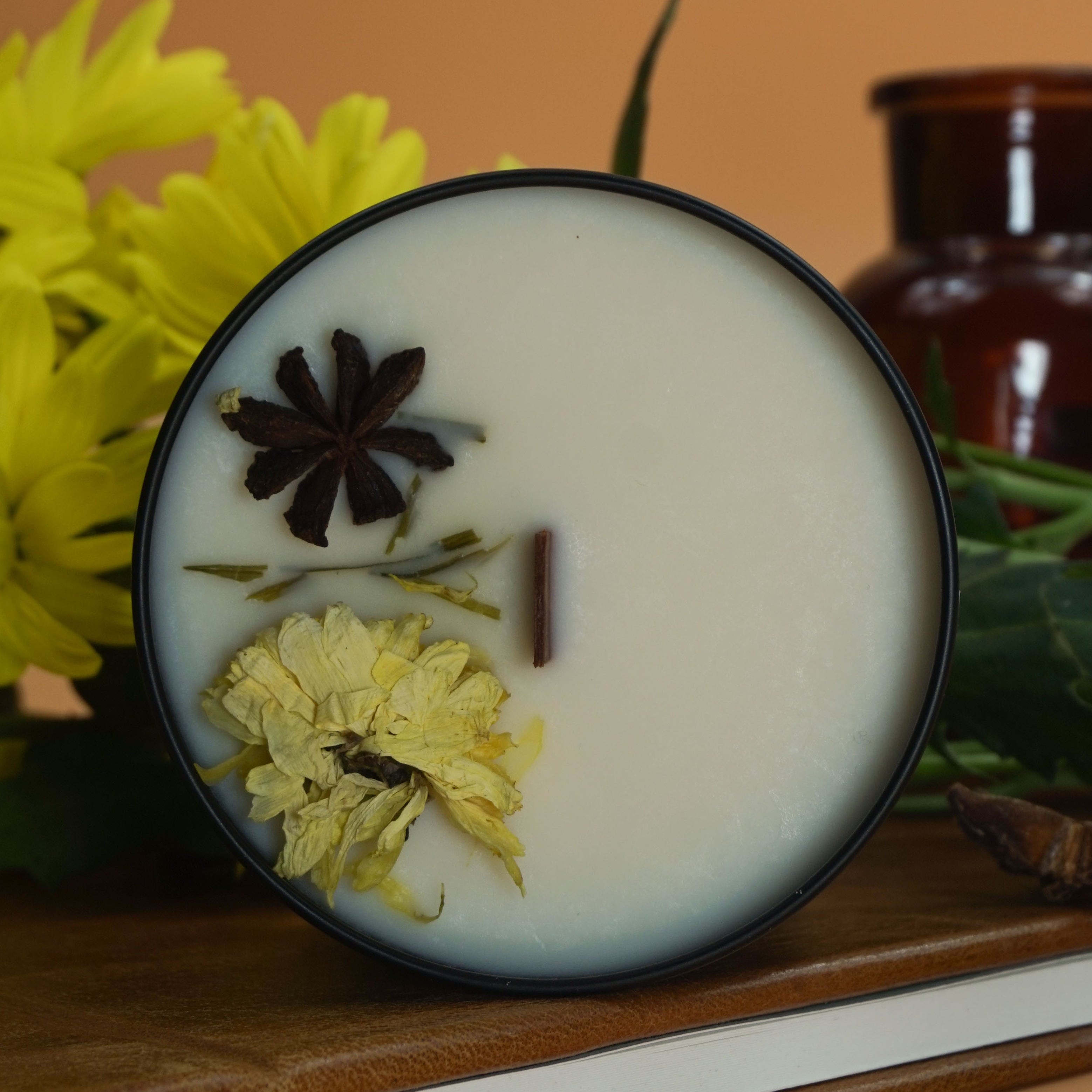 Cette image montre une bougie parfumée dans un pot noir. La bougie est posée sur un carnet en cuir marron avec un bouton-pression doré. Autour de la bougie, il y a des fleurs jaunes, probablement des chrysanthèmes, et des étoiles de badiane (anis étoilé). En arrière-plan, on peut voir une bouteille en verre brun et des feuilles vertes. La bougie a une mèche en bois et est décorée avec des fleurs séchées et des étoiles de badiane sur le