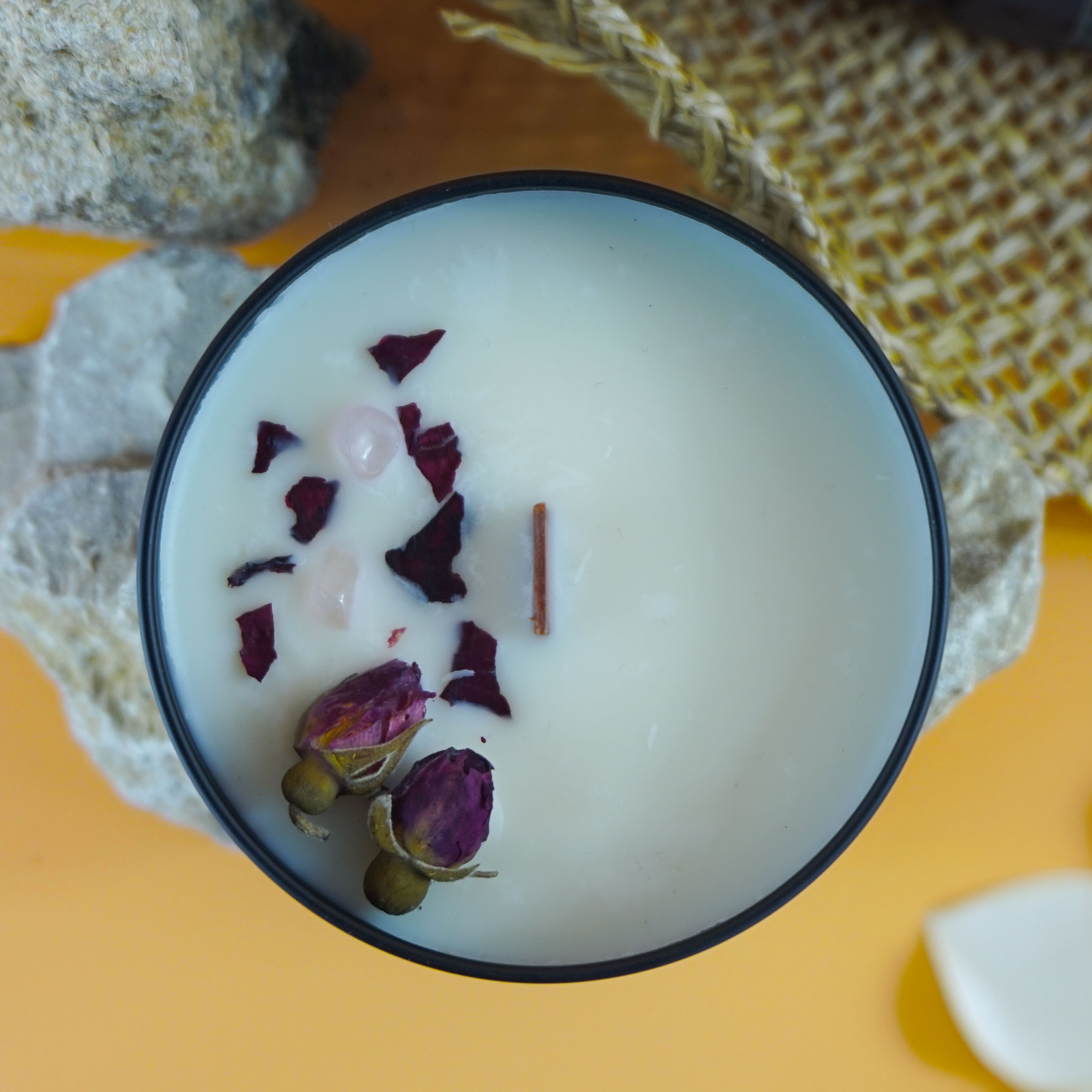 L'image montre une bougie dans un contenant noir.
La bougie est posée sur des pierres, avec un chapeau tissé et des fleurs blanches (probablement des frangipaniers) en arrière-plan. Le fond est d'un orange vif, ce qui contraste avec les éléments naturels au premier plan. L'ensemble de la composition, avec la bougie, les pierres, le chapeau et les fleurs, crée une atmosphère sereine et visuellement attrayante.
