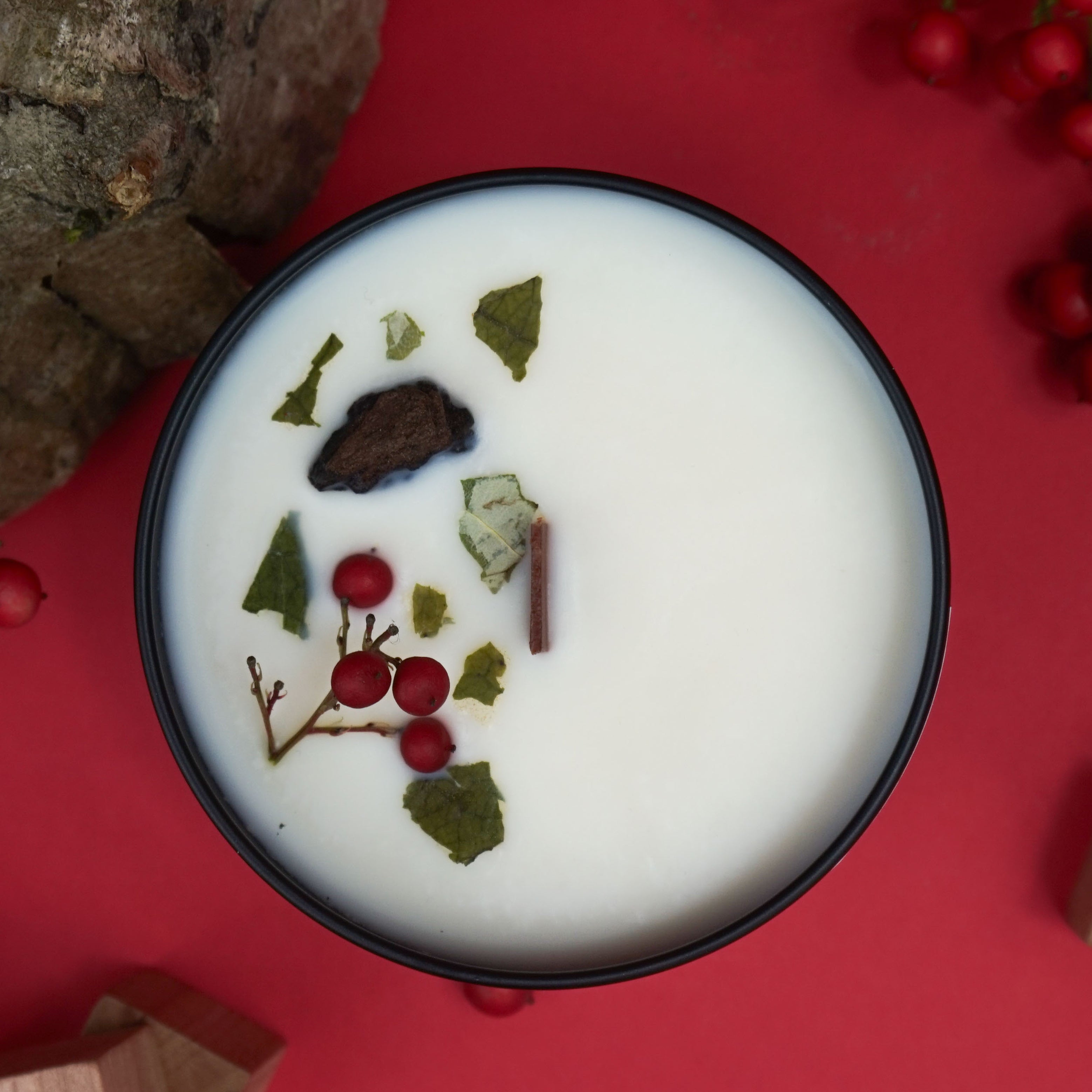 L'image montre une bougie parfumée dans un récipient noir.
La bougie est entourée de divers éléments décoratifs, notamment des baies rouges, des feuilles vertes, des cubes en bois et un morceau d'écorce d'arbre. Le fond est d'un rouge uni, contrastant avec les éléments naturels et la bougie, créant une composition visuellement attrayante.
