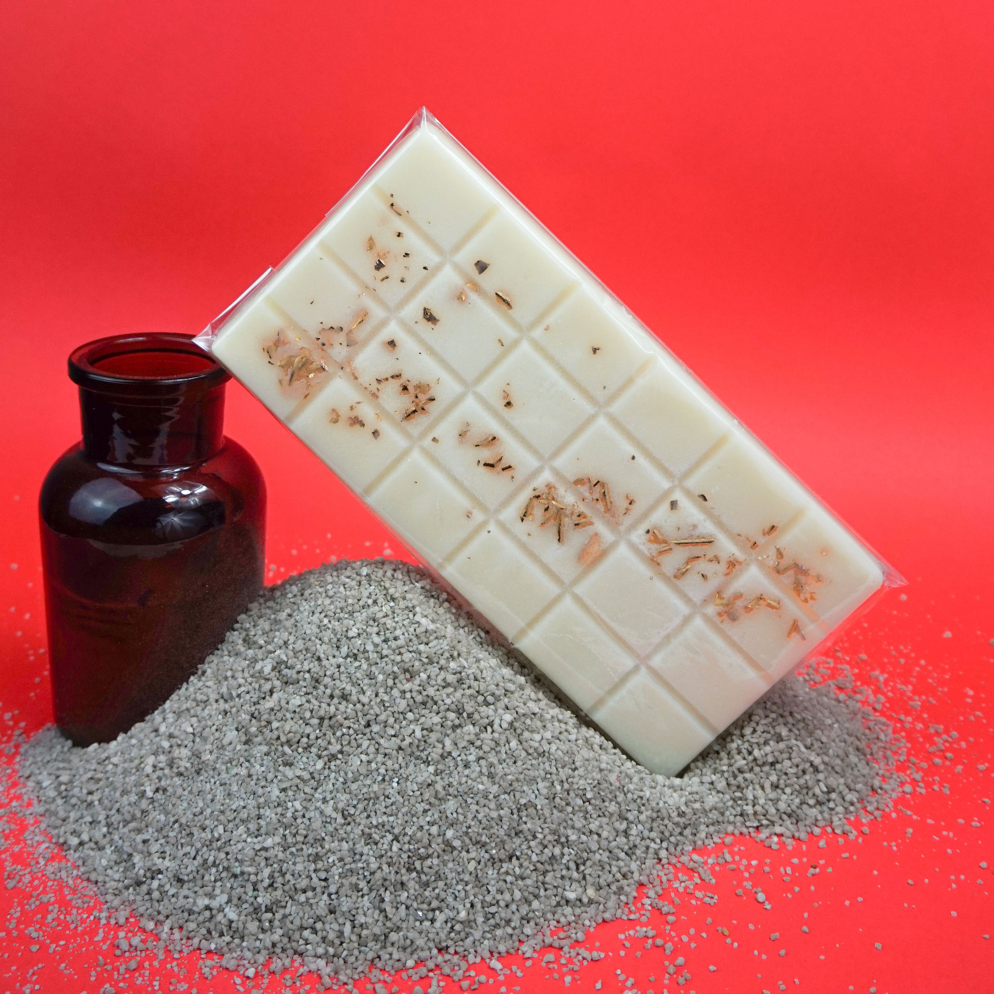 L'image montre une barre rectangulaire blanche avec un motif en grille, ressemblant à une tablette de chocolat mais probablement un fondant de cire ou une barre de savon. La barre a des herbes séchées ou des fleurs intégrées. Elle est appuyée contre un petit tas de matière granuleuse grise, peut-être du sable ou du gravier. À côté du tas se trouve une bouteille en verre brun foncé. Le fond est d'un rouge vif, ce qui fait ressortir les objets.