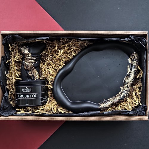 L'image montre une boîte cadeau contenant une bougie noire et un objet décoratif noir, tous deux accentués de doré. La bougie est étiquetée "LUMIÈRE de Genève" et "AMOUR FOU", et est décrite comme "ARTISANAL" et "100% NATUREL". L'objet décoratif a une forme irrégulière avec une surface lisse et des accents dorés sur un côté. Les articles sont nichés dans du papier de remplissage déchiqueté dans une boîte en carton. L'arrière-plan de l'image est une combinaison de formes géométriques rouges et noir.