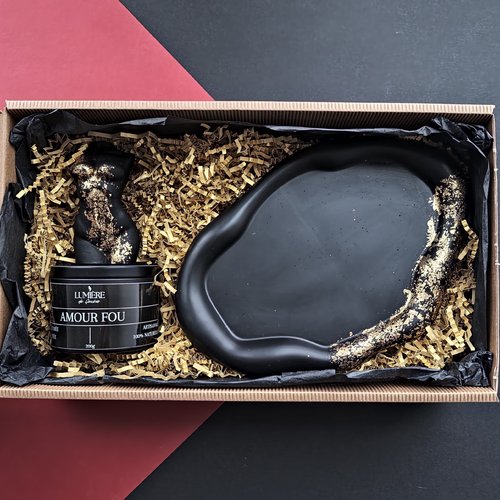 L'image montre une boîte cadeau contenant deux articles : une bougie noire et une assiette décorative noire. La bougie est étiquetée "LUMIÈRE de Genève" et "AMOUR FOU," décrite comme artisanale et 100% naturelle, avec un poids de 200g. Elle a une forme unique et est décorée de flocons dorés. L'assiette décorative est également noire avec une forme irrégulière et ondulée, ornée de flocons dorés sur un côté. Les articles sont nichés dans du papier de remplissage déchiqueté à l'intérieur d'une boîte en carton.