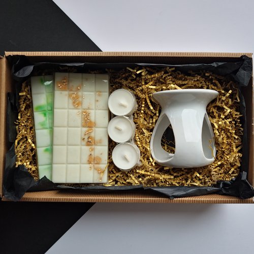 L'image montre une boîte cadeau contenant plusieurs articles disposés sur un lit de papier doré déchiqueté. Les articles comprennent :

Un brûleur d'huiles en céramique blanc.

Trois bougies chauffe-plat blanches.

Deux barres de fondants de cire, l'une avec des accents verts et l'autre avec des flocons dorés.

Cette composition présente un coffret cadeau soigneusement conçu, probablement destiné à l'aromathérapie ou aux parfums d'intérieur. 