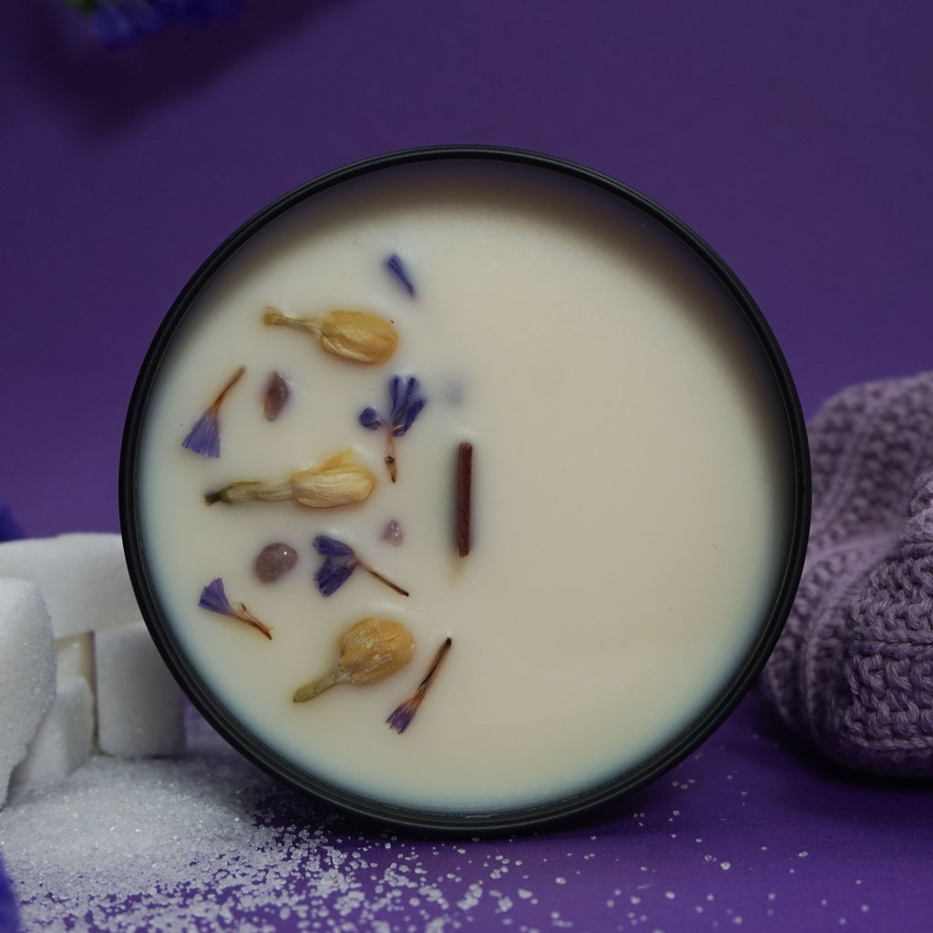 Image d'une bougie parfumée dans un contenant noir. La bougie est décorée de fleurs séchées et de morceaux d'amande sur sa surface. À gauche, on voit des morceaux de sucre et des fleurs violettes. À droite, un tissu en tricot mauve complète la composition, le tout posé sur un fond violet qui renforce une ambiance douce et apaisante