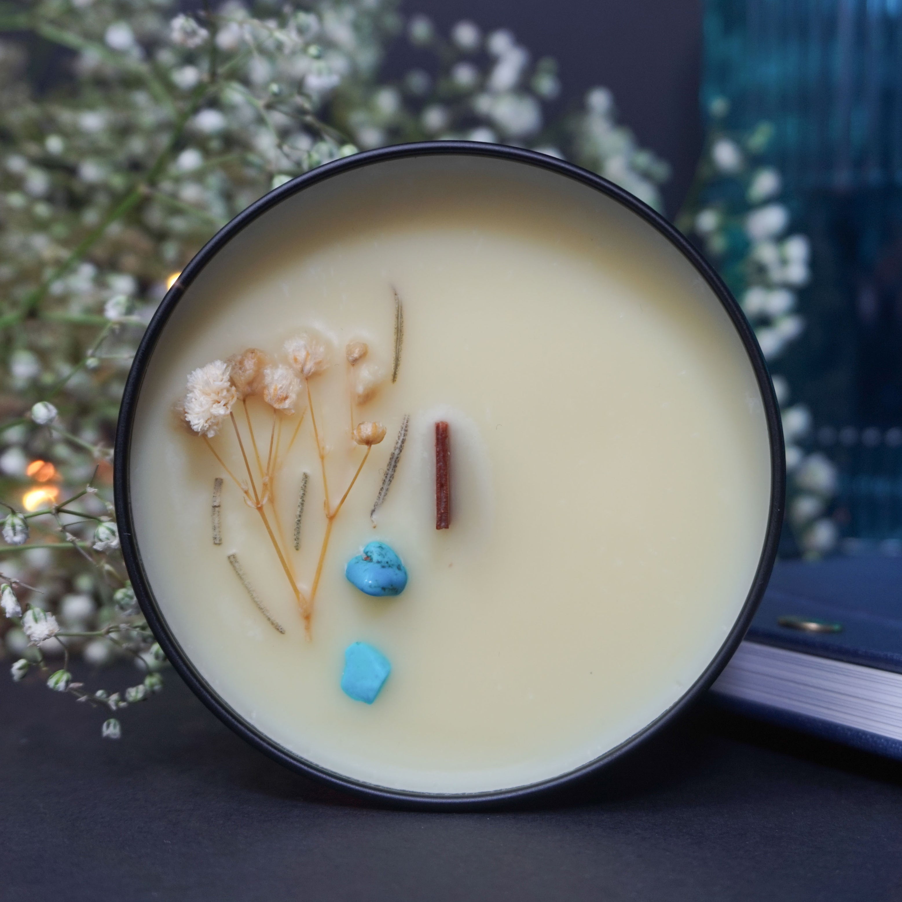 Cette image montre une bougie dans un récipient noir.
La bougie est placée sur une surface bleue, probablement un livre, avec un petit bouton métallique visible. En arrière-plan, on voit de petites fleurs blanches et quelques lumières floues, créant une atmosphère cosy et esthétique. La bougie elle-même a une mèche en bois et des éléments décoratifs, des fleurs séchées et une petite pierre bleue incrustée dans la cire.