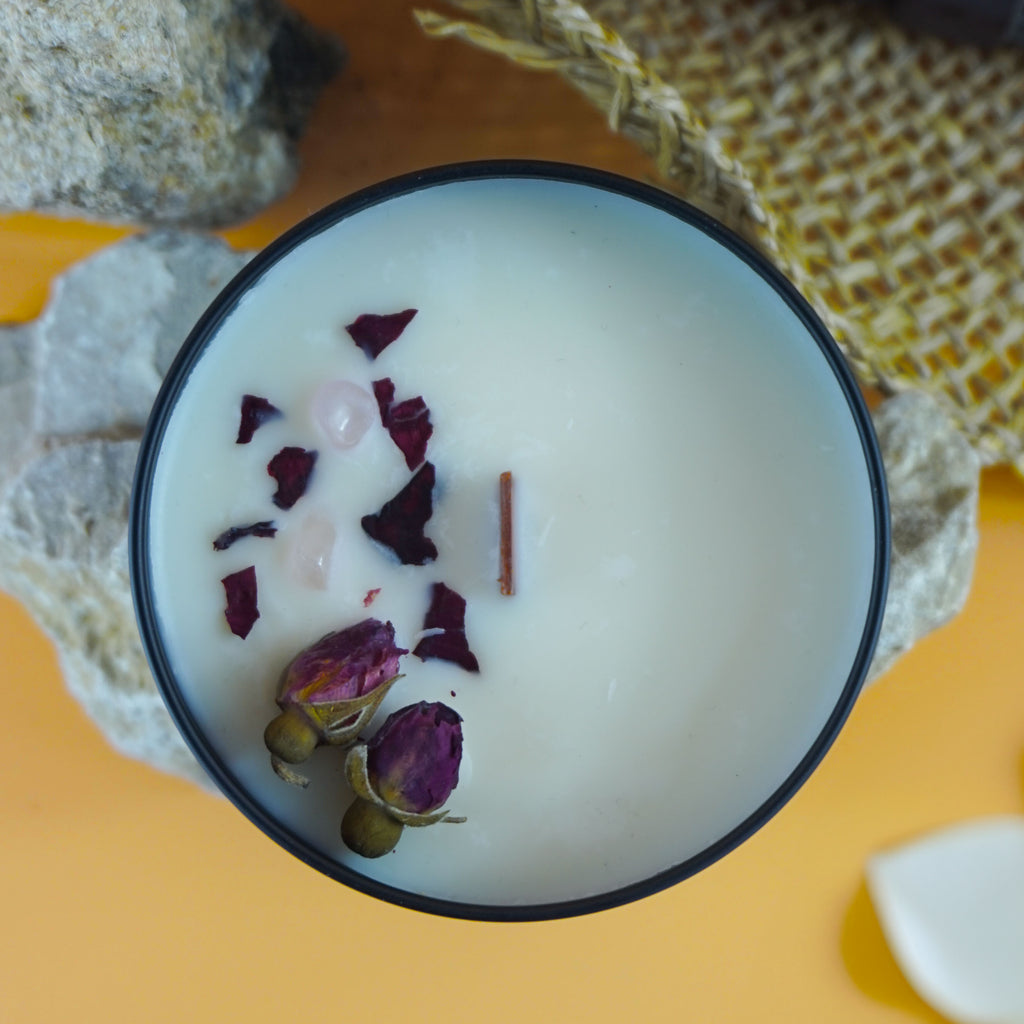 L'image montre une bougie dans un contenant noir.
La bougie est posée sur des pierres, avec un chapeau tissé et des fleurs blanches (probablement des frangipaniers) en arrière-plan. Le fond est d'un orange vif, ce qui contraste avec les éléments naturels au premier plan. L'ensemble de la composition, avec la bougie, les pierres, le chapeau et les fleurs, crée une atmosphère sereine et visuellement attrayante.