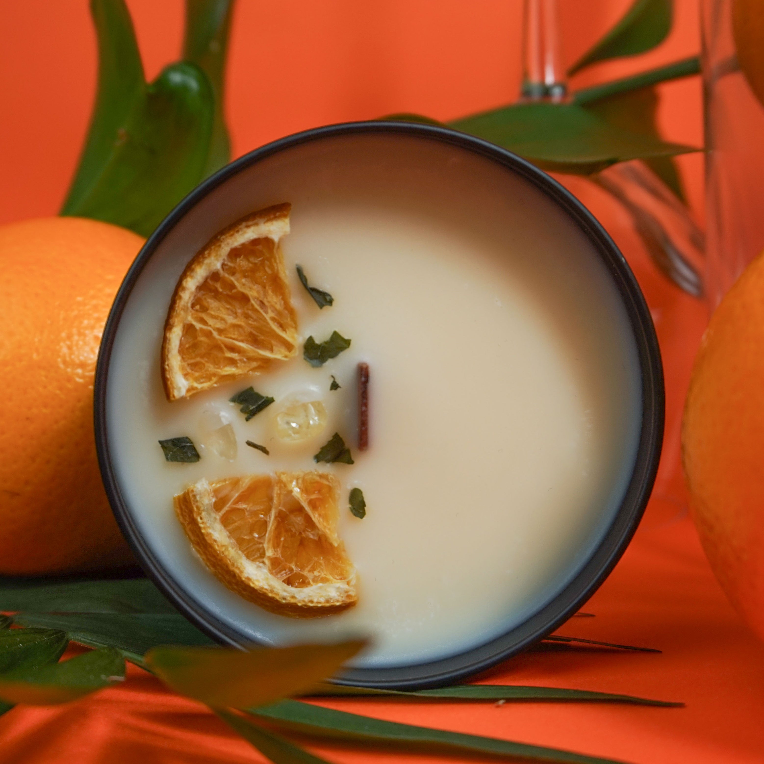 Photographie d'une bougie parfumée dans un contenant noir .
La bougie est décorée de morceaux séchés d'orange et de feuilles sur sa surface. Elle est entourée de grandes oranges fraîches et de feuilles vertes, avec un fond orange vif qui évoque une ambiance fraîche et vivifiante