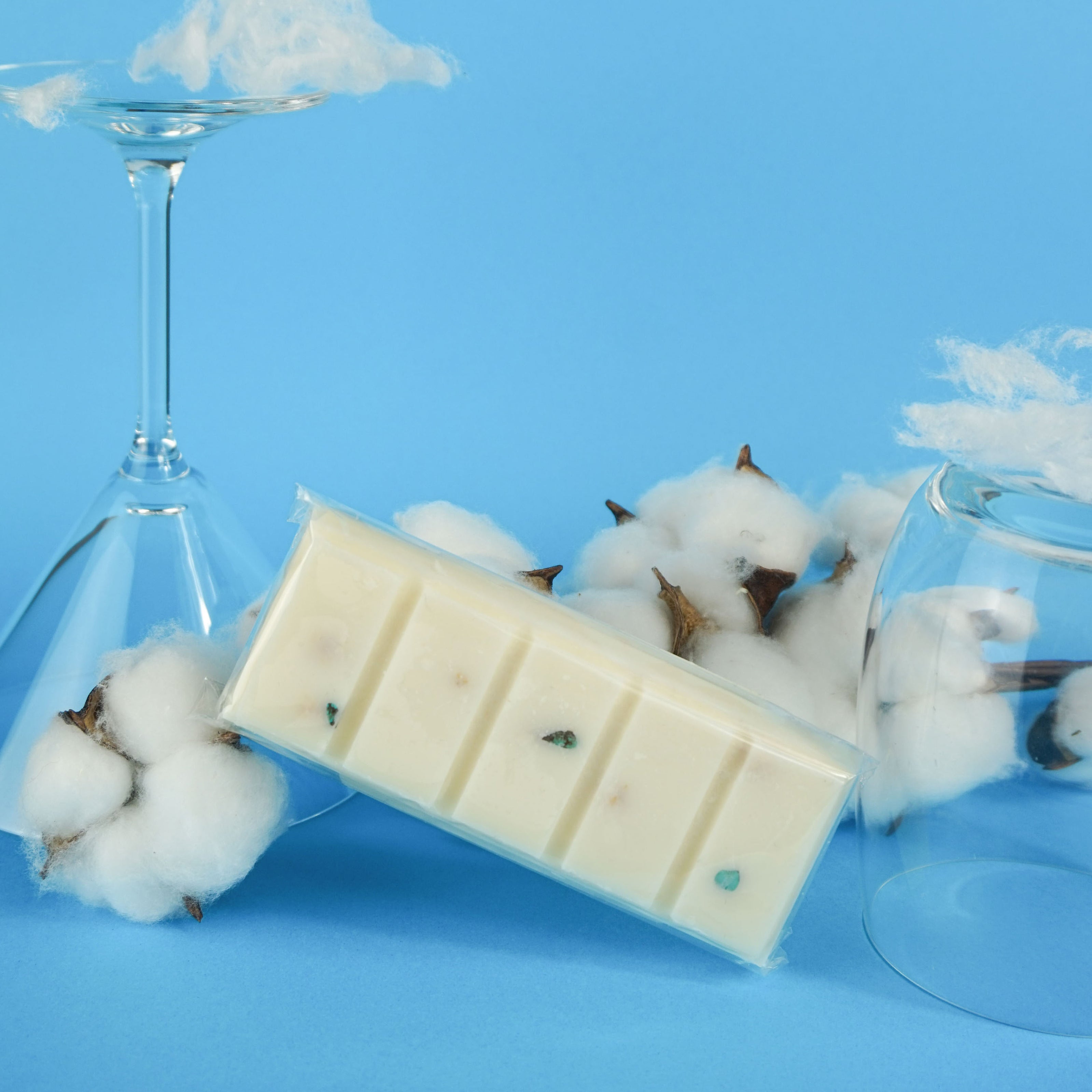 L'image montre une tablette de chocolat blanc avec de petites inclusions vertes, posée sur un fond bleu. La tablette est segmentée en cinq morceaux et est enveloppée dans du plastique transparent. Autour de la tablette de chocolat, on voit des capsules de coton et deux verres à vin retournés, l'un d'eux contenant du coton à l'intérieur et sur le dessus. La composition de l'image est intéressante visuellement en raison du contraste entre le chocolat blanc et le fond bleu.