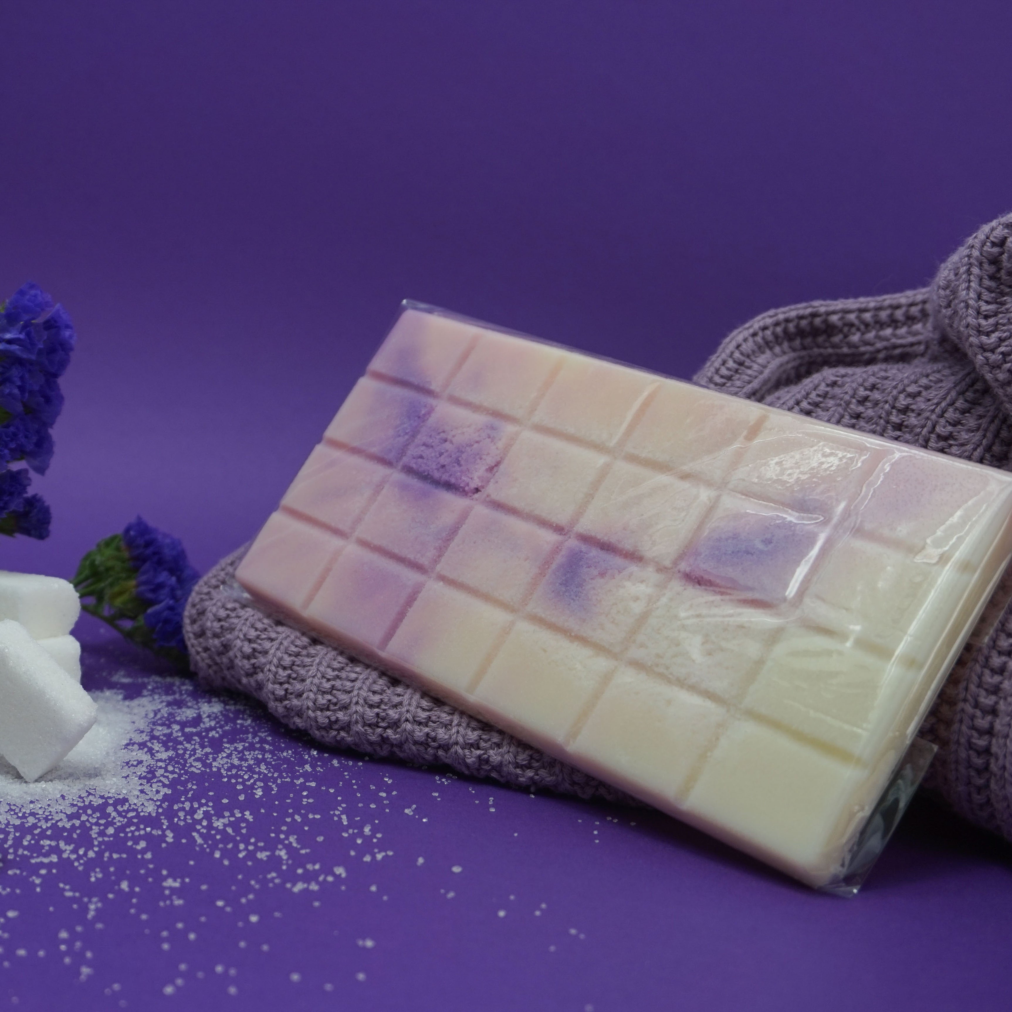 L'image montre une barre de savon avec un motif en grille, présentant un dégradé de couleurs allant du blanc au violet. Le savon est placé sur un tissu tricoté, avec des granulés blancs éparpillés autour, représentant possiblement des sels de bain. Il y a aussi des fleurs violettes et des cubes de sucre blanc en arrière-plan, le tout sur un fond violet. La combinaison des éléments et des couleurs crée une composition visuellement attrayante et apaisante.