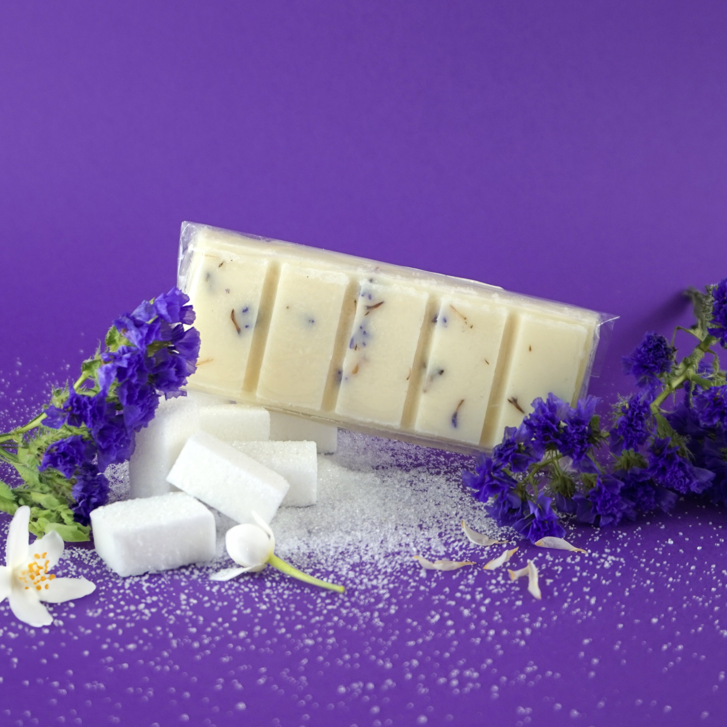 L'image montre des fondants de cire ou barres de savon blancs avec des inclusions violettes et brunes, emballés dans du plastique transparent. Ils sont entourés de cubes de sucre blanc et de fleurs violettes, sur un fond violet uni. Les éléments créent un contraste visuellement attrayant. 