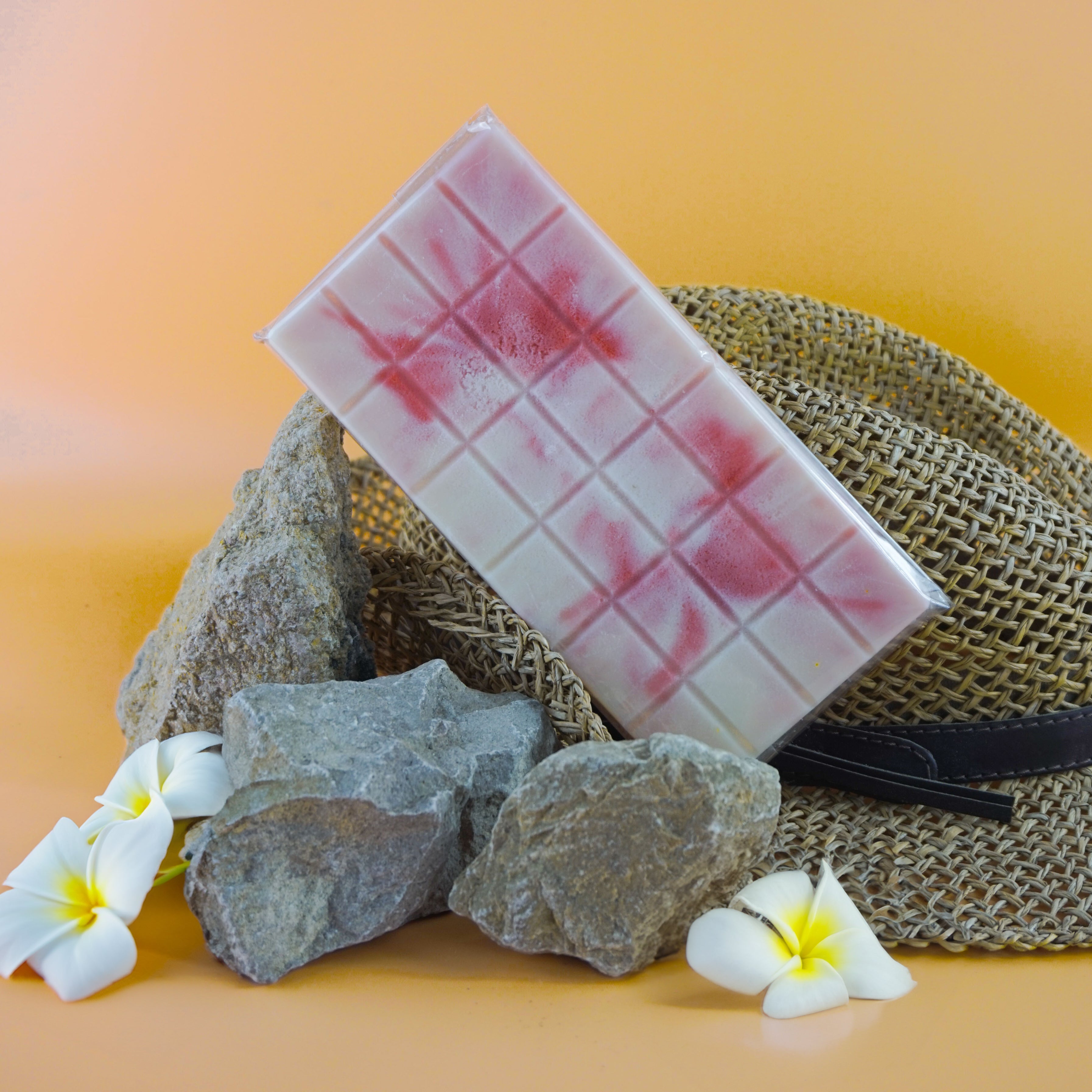 L'image montre une barre rectangulaire avec un motif en grille, présentant un mélange de couleurs blanches et rouge-rosé. La barre est placée contre un fond de roches, de fleurs blanches et d'un chapeau tissé avec un ruban noir, le tout sur un fond orange clair. La combinaison des éléments naturels comme les roches et les fleurs avec la barre et le chapeau crée une composition visuellement attrayante.