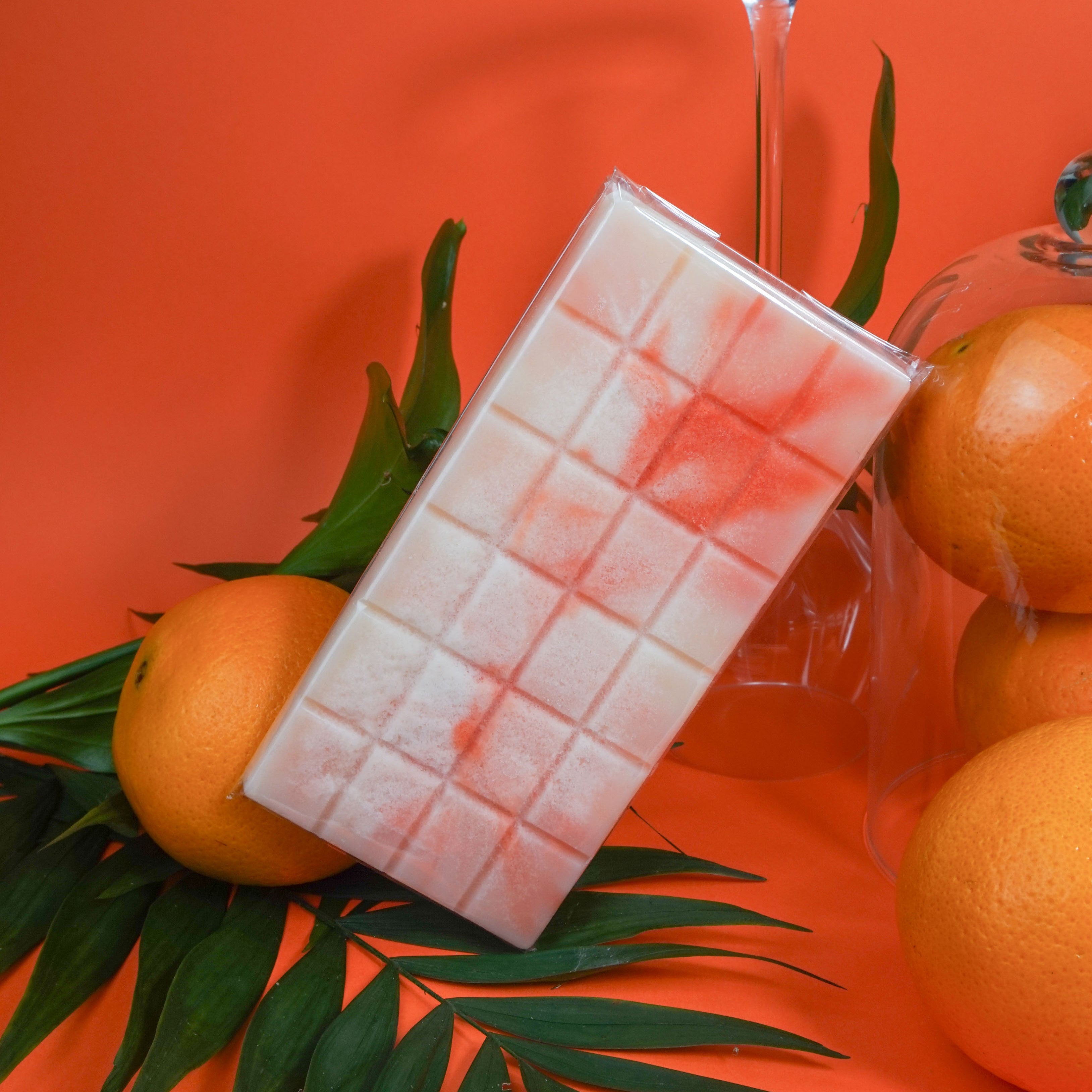 L'image montre une barre de chocolat blanc avec un motif en grille, avec des traînées rouges-orangées, appuyée contre un fruit orange. Le fond est d'un orange uni, et il y a des feuilles vertes et d'autres oranges autour de la barre de chocolat. L'ensemble est visuellement attrayant grâce aux couleurs contrastantes et au thème tropical créé par les feuilles et les oranges.