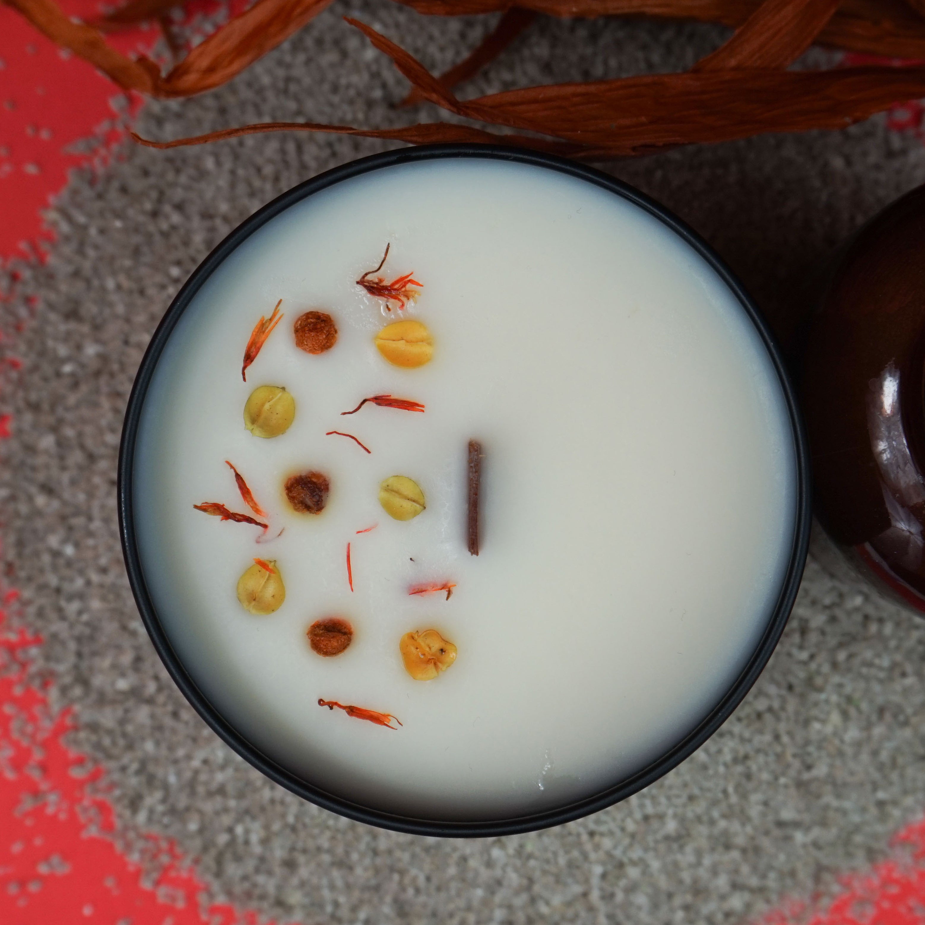 L'image montre une bougie parfumée dans un récipient noir.
La bougie est posée sur un lit de petits galets blancs, à côté d'une bouteille en verre brun foncé. En arrière-plan, il y a un bouquet de feuilles séchées brunes. Le fond est d'une couleur rouge vif, ce qui contraste avec les tons terreux de la bougie, de la bouteille et des feuilles. La bougie elle-même a des petits morceaux de matière séchée sur sa surface.