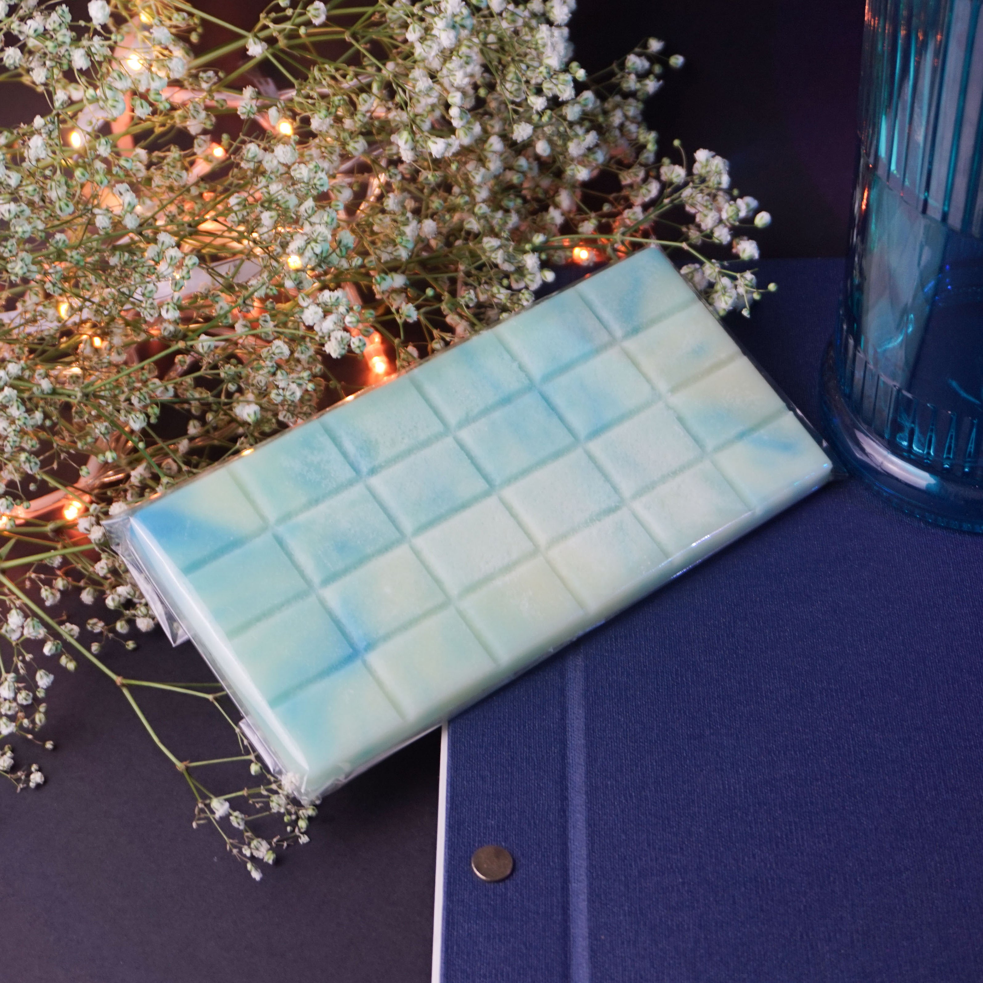 L'image montre une barre rectangulaire, probablement du chocolat ou une confiserie, avec un motif de grille sur sa surface. La barre a un dégradé de couleur allant du bleu clair au jaune pâle. Elle est posée sur une surface bleue foncée, à côté d'un objet cylindrique bleu, peut-être un vase ou un contenant. Autour de la barre se trouvent de délicates fleurs blanches et de petites lumières féeriques chaudes, créant une atmosphère visuellement attrayante et cosy.